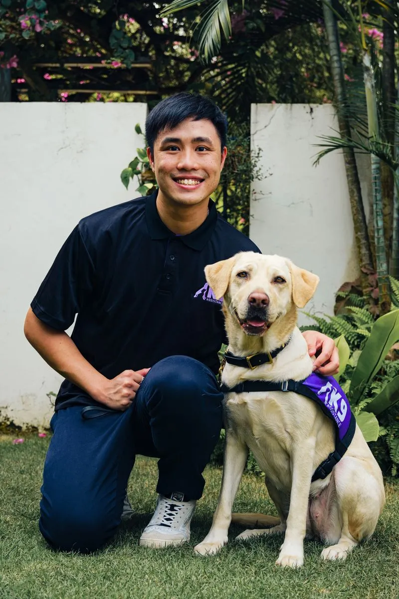 Mr Vincent Lim, K9Assistance Communications and Community Partnerships Executive, knelt down next to a golden Labrador Retriever wearing a purple K9Assistance vest. 