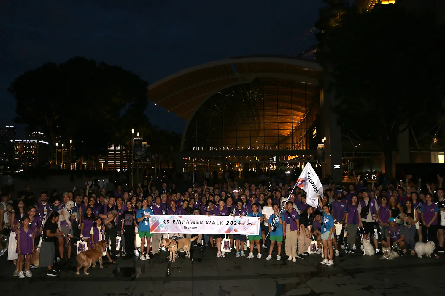 A large group photo taken at K9 Empawer Walk 2024, at night. A banner being held up at the front reads "K9 Empawer Walk 2024". Participants are wearing purple vests, and some have their dogs with them.