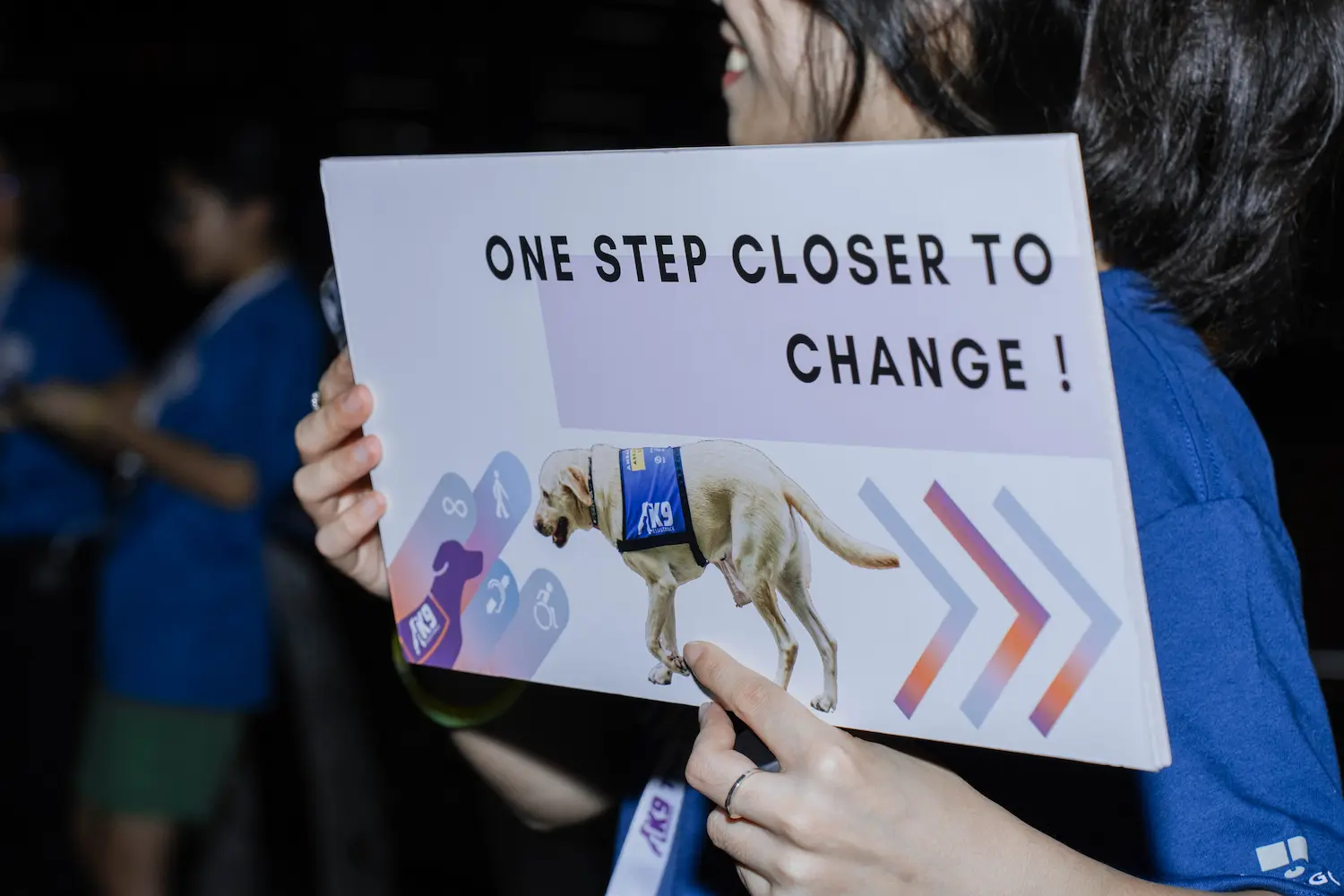 Closeup of a directional signage with graphics of an Assistance Dog and arrows, held up by a smiling volunteer. Text on the sign reads: 'One step closer to change!'