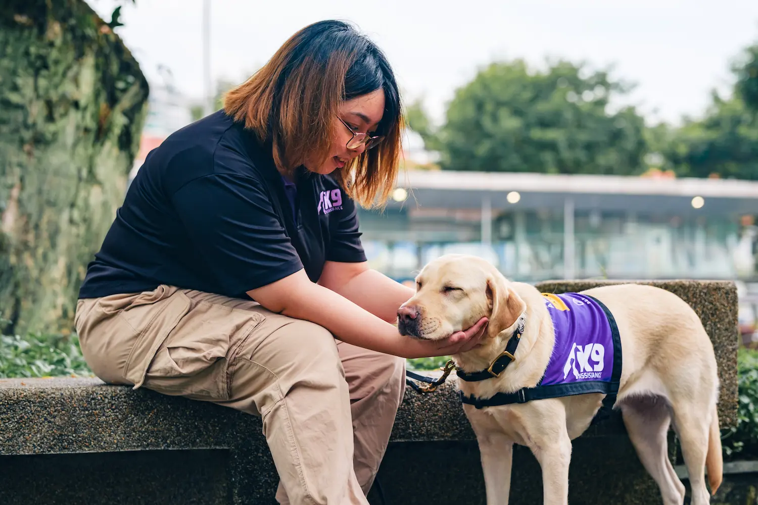 K9Assistance's Ambassador Assistance Dog standing next to its seated handler. The dog is resting its head on the handler's hand under its chin, while the handler looks at the dog.